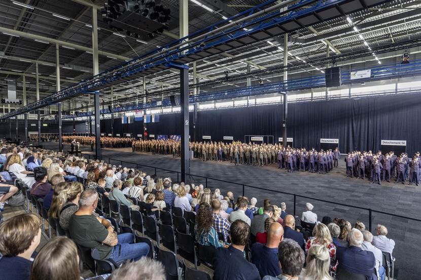 750 militairen staan opgesteld in een grote hal, publiek op de tribune kijkt toe.