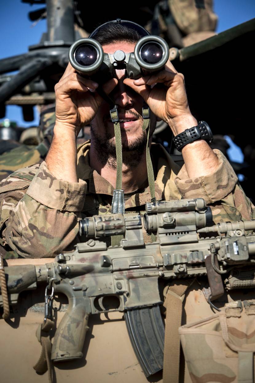 Een commando met een HK416A5-geweer om zijn nek observeert zijn omgeving met een verrekijker.