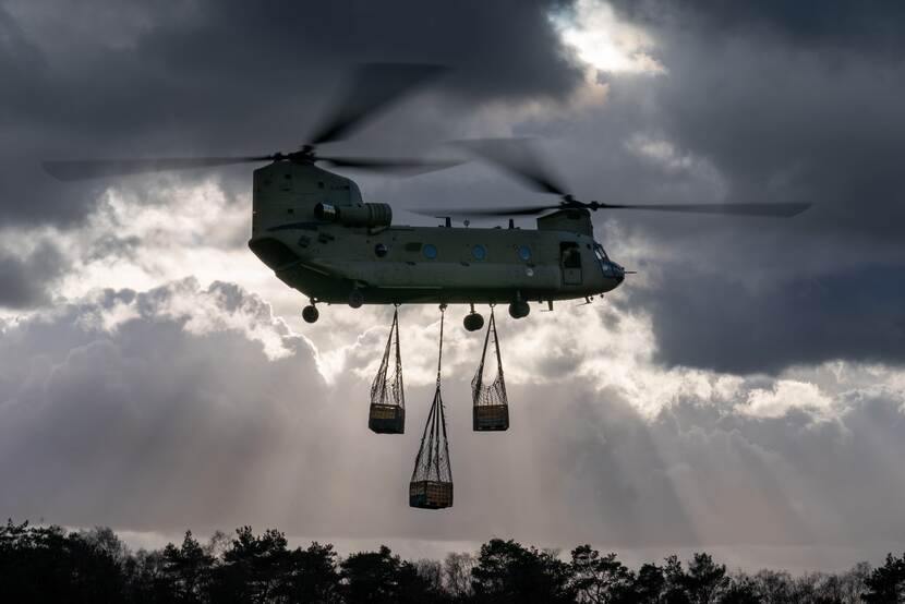Een Chinook-transporthelikopter in de lucht, er hangen 3 netten met houten kisten aan de onderkant.