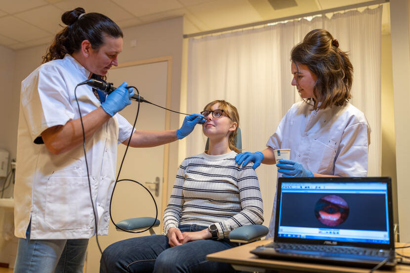 2 verpleegkundigen in witte doktersjassen brengen een flexibele scoop met camera in bij een mevrouw via de neus. Rechts op tafel staat een laptop waarop de beelden van de camera te zien zijn.