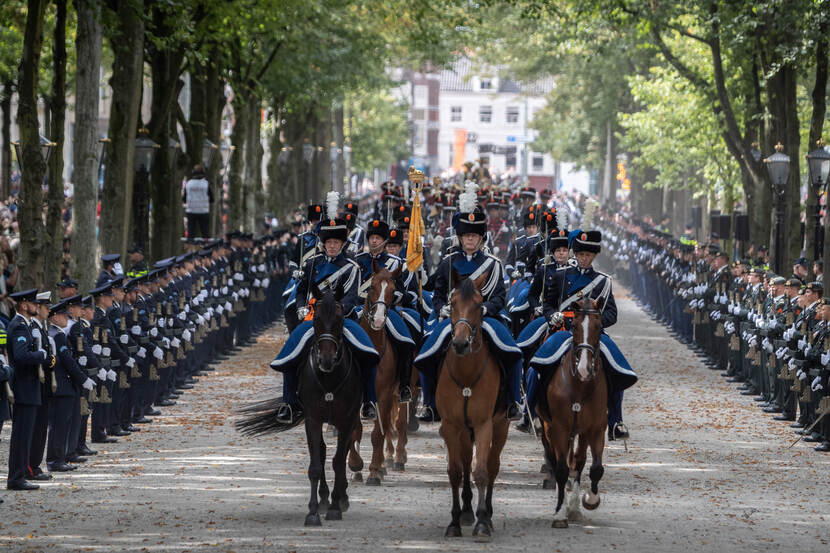 Marechaussees in ceremonieel tenue op paarden. Links en rechts langs de weg een ere-afzetting.