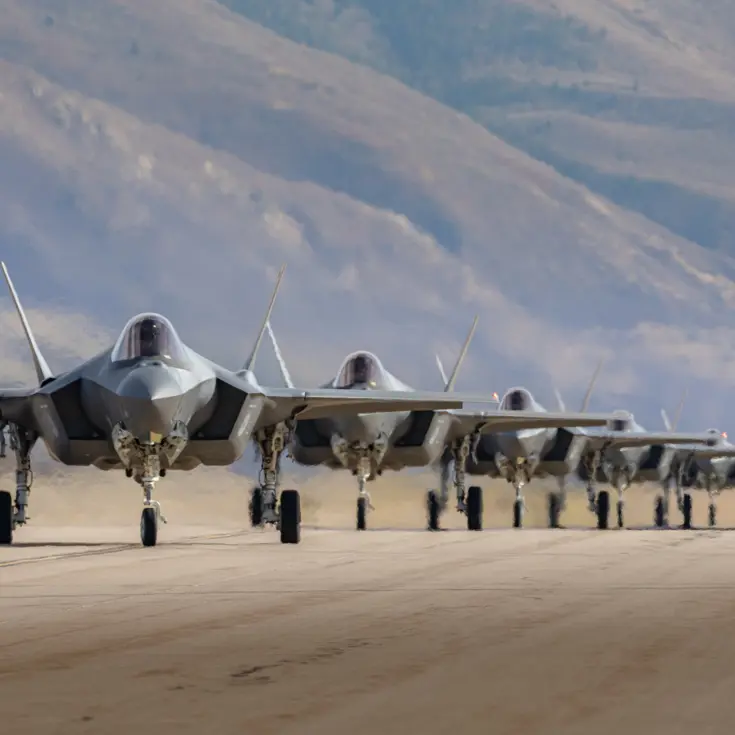 Vijf F-35’s taxiën achter elkaar op een startbaan met op de achtergrond bergen.
