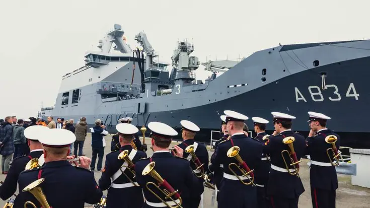Marineschip ligt langs kade. Op voorgrond militaire muziekkapel.