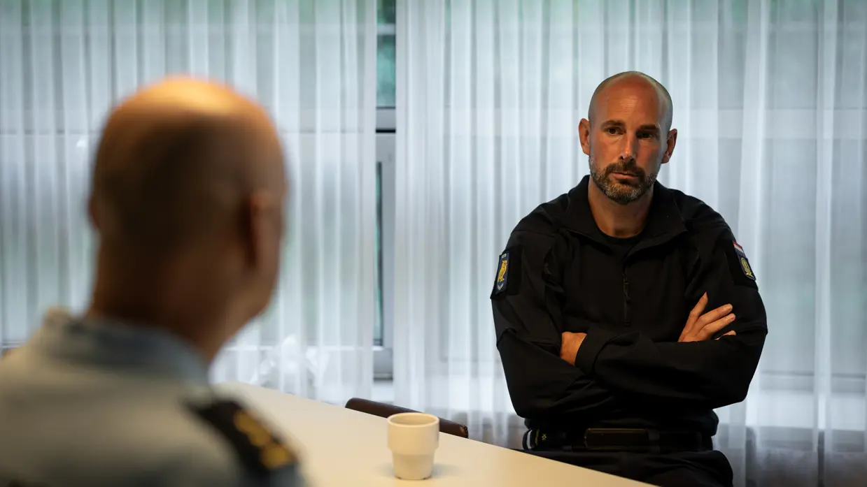 Majoor Arno zit aan een tafel terwijl hij luistert naar overste David.
