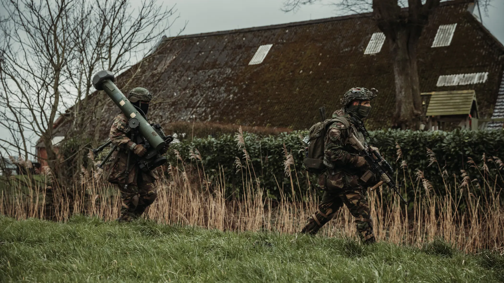 Twee gewapende militairen lopen achter elkaar door een grasveld. De achterste draagt een cilindervormig, donkergroen antitankwapen op zijn schouder.