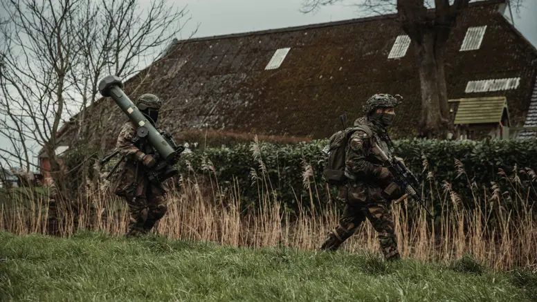 Twee gewapende militairen lopen achter elkaar door een grasveld. De achterste draagt een cilindervormig, donkergroen antitankwapen op zijn schouder.