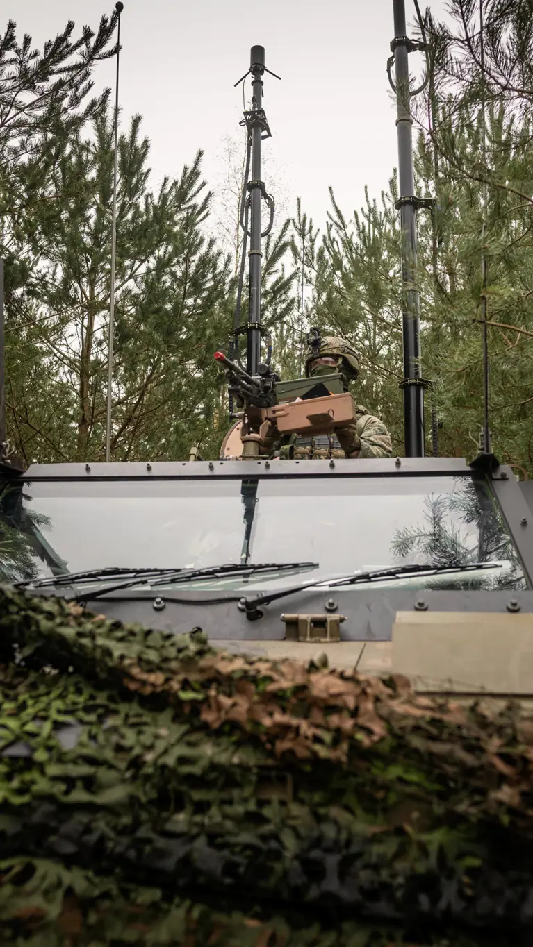 Een militair zit met zijn  halve lichaam bovenop de Bushmaster. Het voertuig is bedekt met een camouflagenet en staat in bosschage. Achter de militair zijn diverse antennes te zien.
