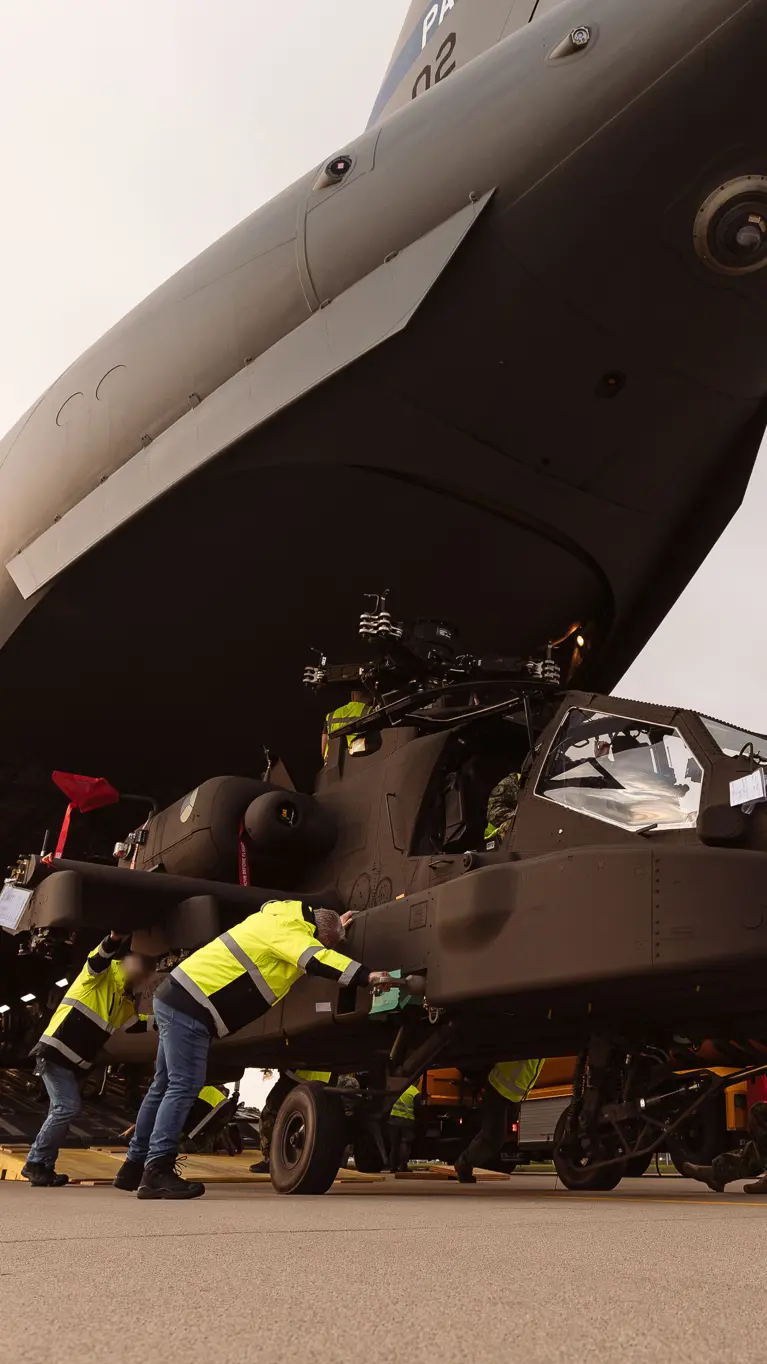 Mannen in gele jassen duwen een Apache uit het laadruim van een groot transportvliegtuig.