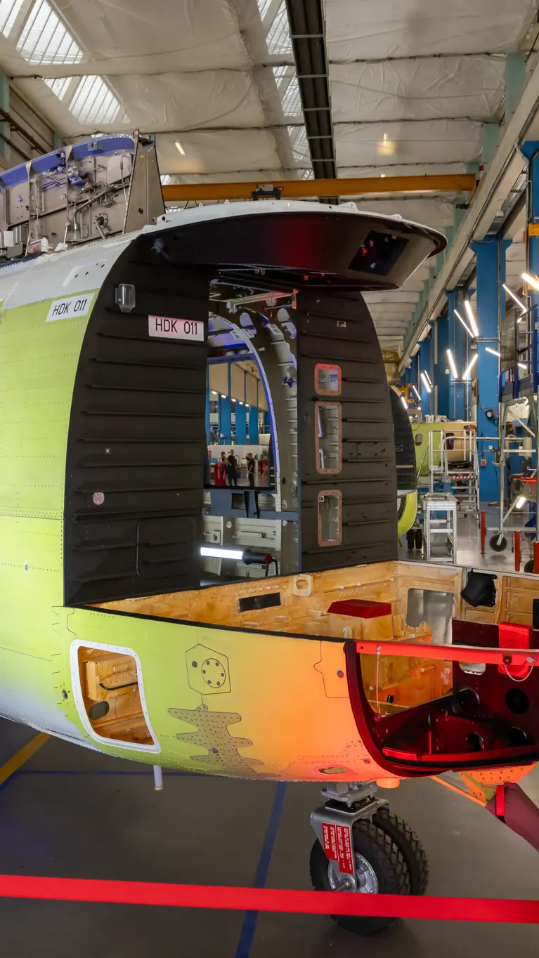 Het nog kale frame van een helikopter in een fabriekshal. Op de voorgrond de Nederlandse en Franse vlag.