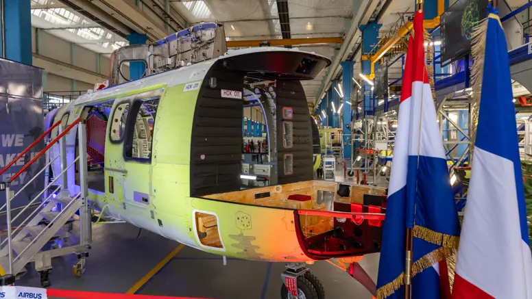 Het nog kale frame van een helikopter in een fabriekshal. Op de voorgrond de Nederlandse en Franse vlag.