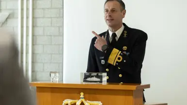 Een sprekende vice-admiraal Jan Willem Hartman, daarnaast zit hij met een burger en een andere militair achter en tafel en zetten zij gelijktijdig een handtekening onder een document.