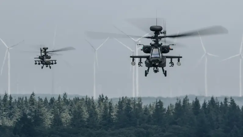 Twee Apaches vliegen boven een bos, op de achtergrond zijn windmolens te zien.