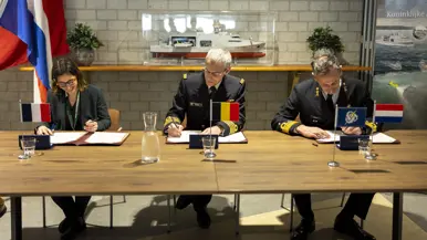 Een sprekende vice-admiraal Jan Willem Hartman, daarnaast zit hij met een burger en een andere militair achter en tafel en zetten zij gelijktijdig een handtekening onder een document.