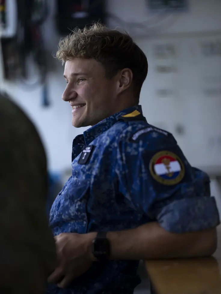 Een lachende, blonde man in blauw uniform vanaf de zijkant gezien. Hij zit op een bankje.