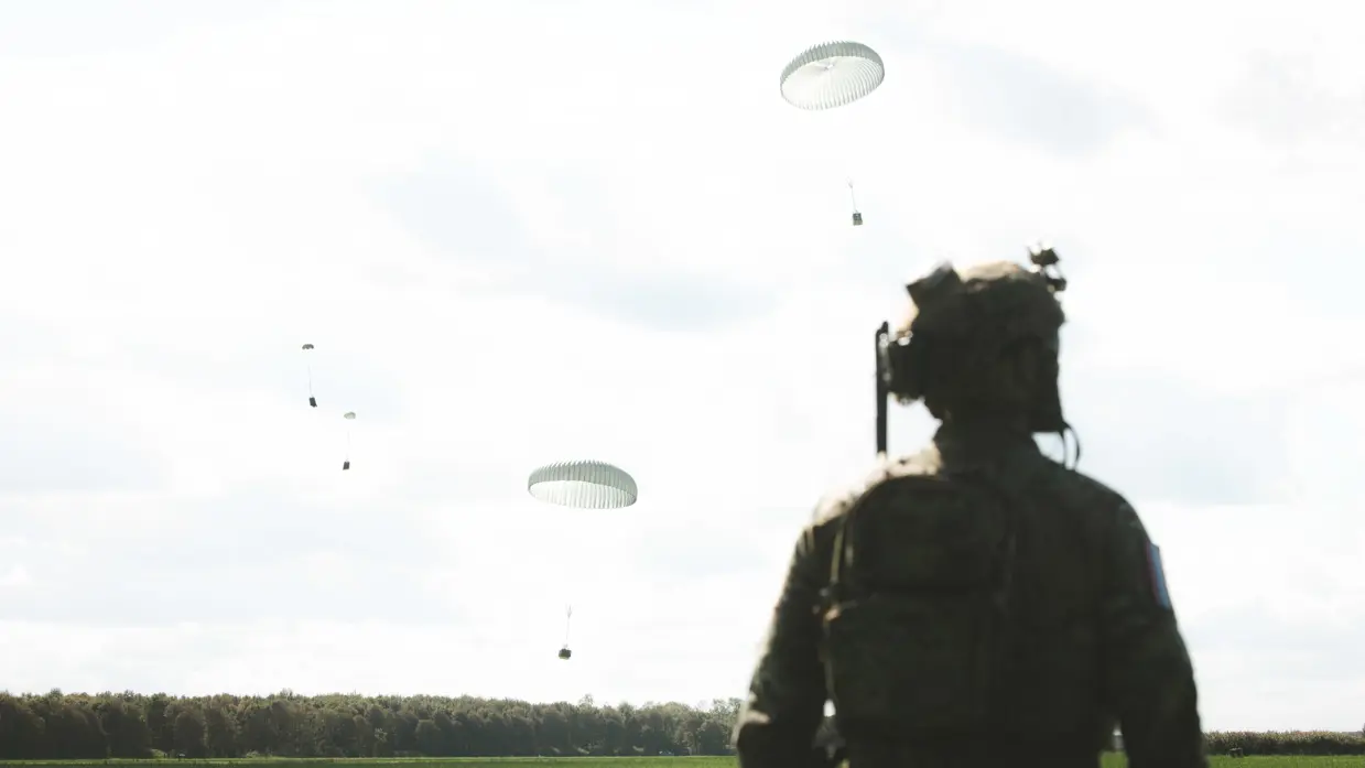 Vanachter gezien staart een militair met helm over een groen veld naar dalende witte parachutes onder licht bewolkte lucht.