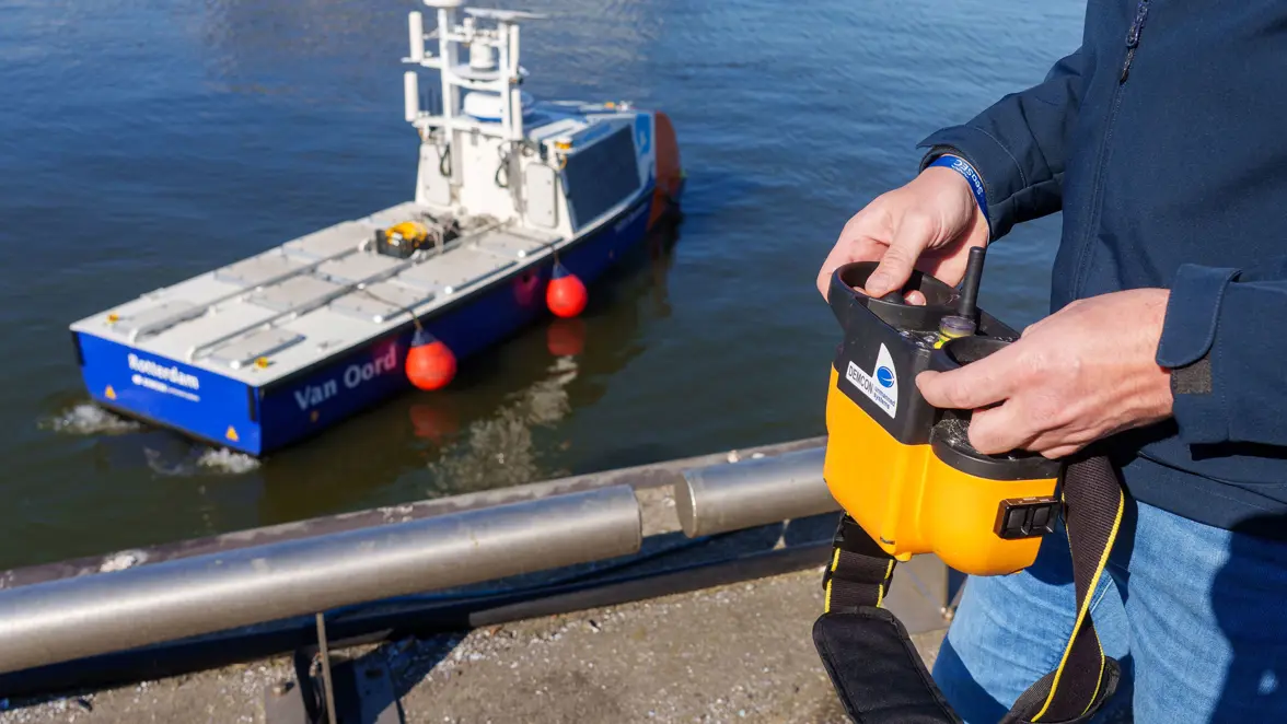 Twee handen besturen via een gele controller een klein bootje met sensoren. 