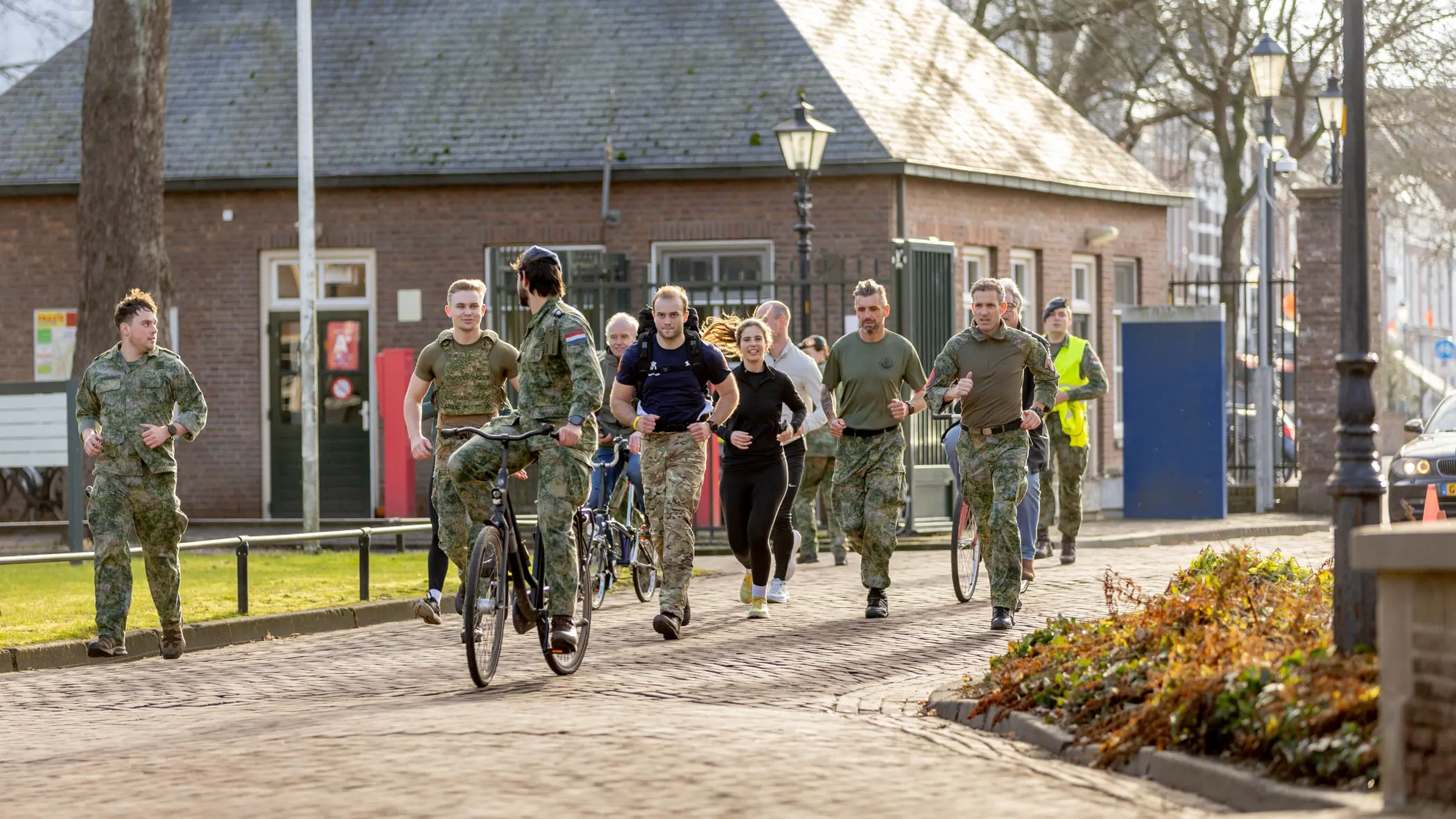 Een groep mensen in beweging op een zonnig pad: sommigen lopen, anderen fietsen. Ze dragen sportieve kleding en militaire uniformen, met bomen en een gebouw op de achtergrond.