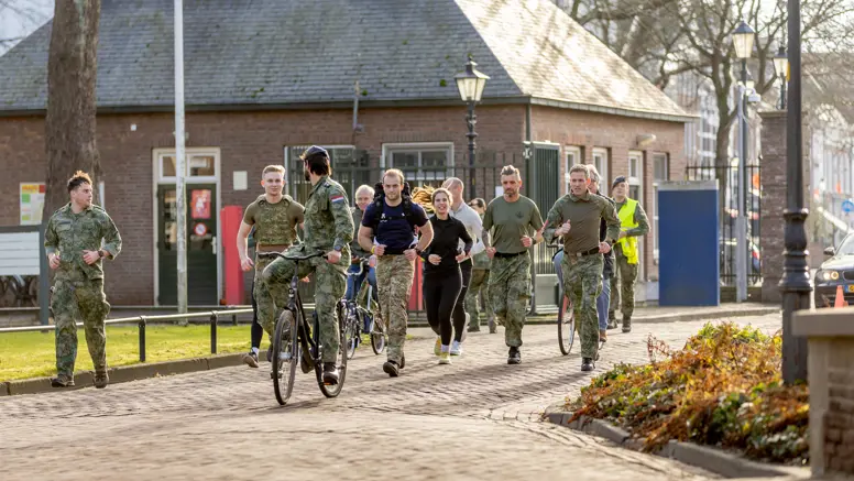 Een groep mensen in beweging op een zonnig pad: sommigen lopen, anderen fietsen. Ze dragen sportieve kleding en militaire uniformen, met bomen en een gebouw op de achtergrond.