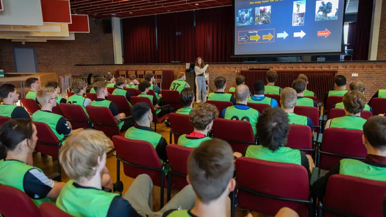 In een zaal houdt een vrouw een voordracht over de luchtmobiele brigade. Studenten in gele vesten luisteren aandachtig naar de presentatie op een groot scherm.&nbsp;