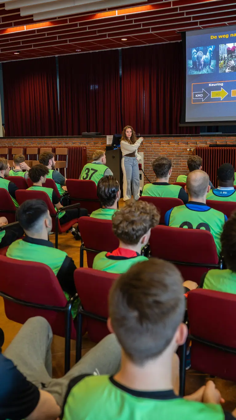 In een zaal houdt een vrouw een voordracht over de luchtmobiele brigade. Studenten in gele vesten luisteren aandachtig naar de presentatie op een groot scherm.&nbsp;
