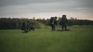 Vijf militairen in een veld met rugzakken en opgevouwen parachutes voor zich.