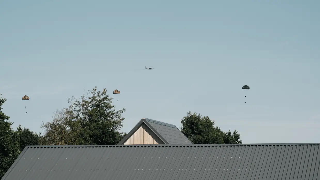 Een grote ronde parachute met camouflagepatroon in tan, bruin en wit hangt volledig open tegen een bewolkte lucht, met twee personen eronder.