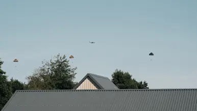 Een grote ronde parachute met camouflagepatroon in tan, bruin en wit hangt volledig open tegen een bewolkte lucht, met twee personen eronder.