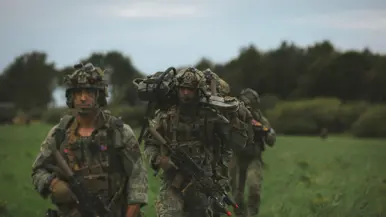 Een rij militairen met helm, wapen en rugzakken lopen door een veld.