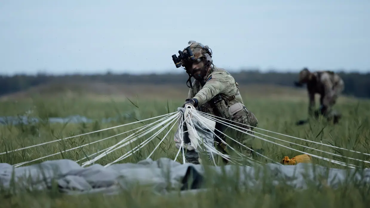 In een grasveld hurkt een militair in camouflage en helm, bundelend witte parachuteleidingen van lichte stof. Op de achtergrond nog een soldaat, onscherp.