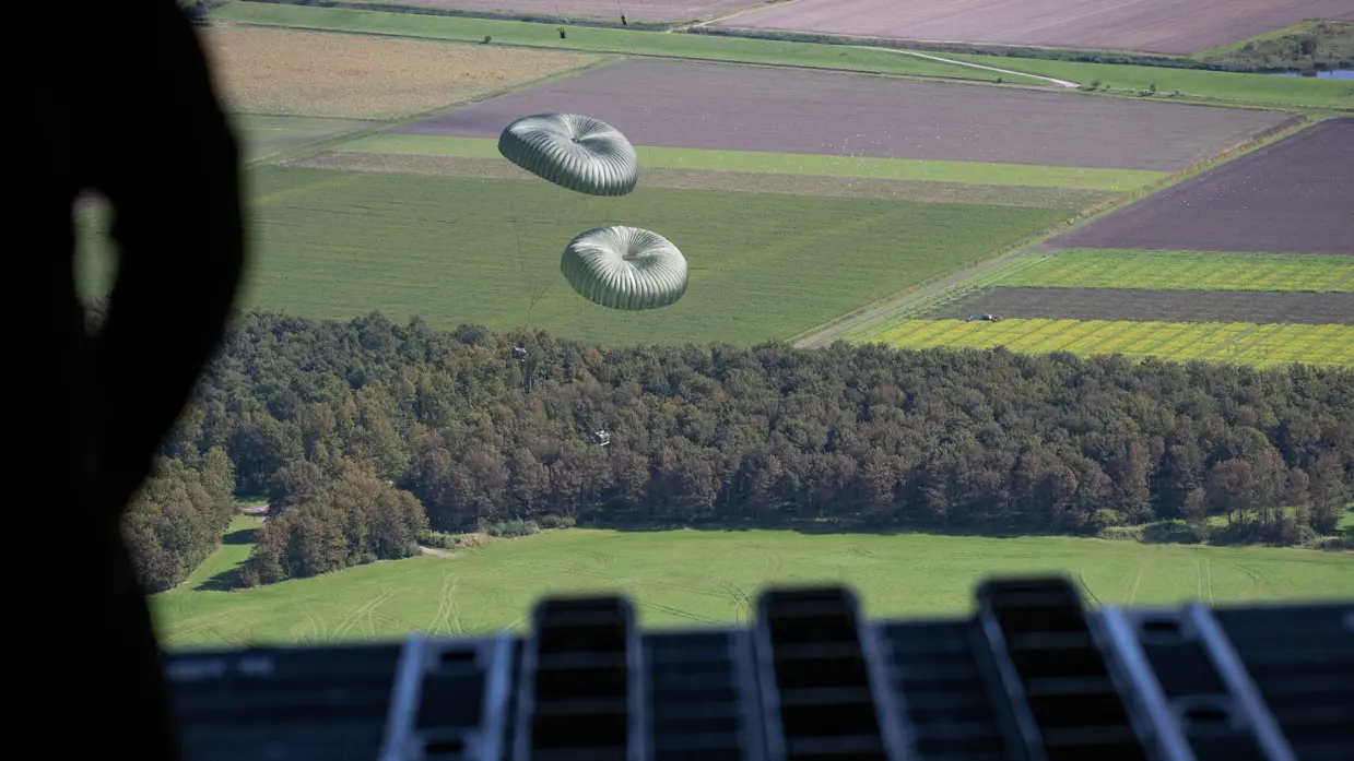 Vanuit de achterzijde van een vliegtuig kijkt een man met een helm naar 2 pakketten aan een parachute.