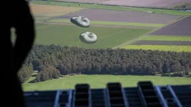 Vanuit de achterzijde van een vliegtuig kijkt een man met een helm naar 2 pakketten aan een parachute.