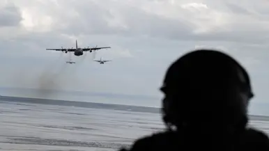 Vanachter een man met een helm zijn twee vliegtuigen zichtbaar, vliegend boven water.