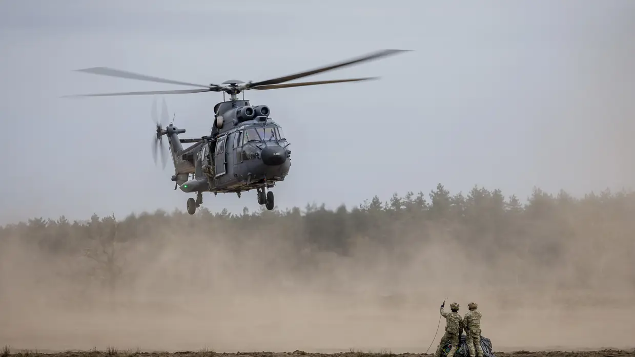 Een militaire helikopter landt in een stoffige, open vlakte. Twee soldaten staan op de grond en wijzen naar de helikopter. 