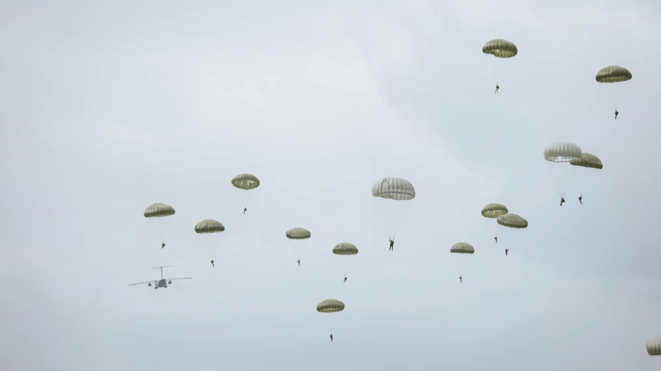 Para-chutisten dalen neer onder groene parachutes; op de achtergrond vliegt een grijs militair transportvliegtuig van links naar rechts onder een bewolkte, grijze lucht.