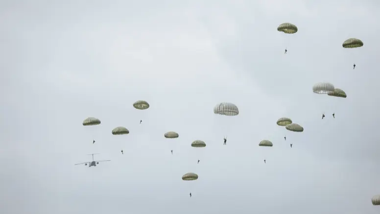 Para-chutisten dalen neer onder groene parachutes; op de achtergrond vliegt een grijs militair transportvliegtuig van links naar rechts onder een bewolkte, grijze lucht.