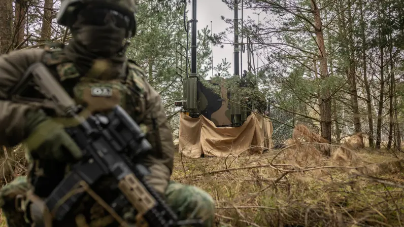 Een gecamoufleerde militair houdt op een knie gewapend de wacht aan de achterzijde van een Bushmaster, die in een groene strook van bos en struik geparkeerd staat. Op de Bushmaster zijn diverse antennes te zien. Foto rechts: Niels kijkt recht in de camera. Hij draagt een lichtgroene blouse en heeft kort donker haar. 