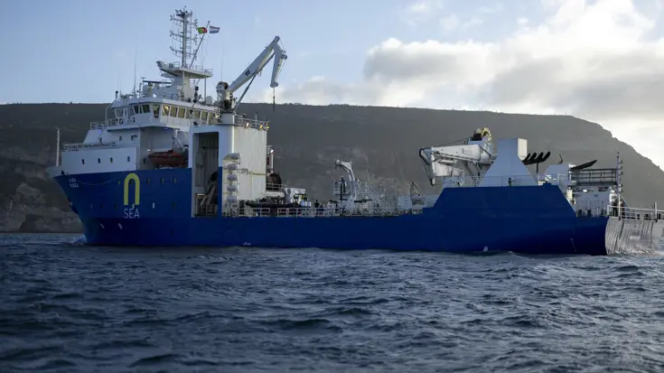Een blauw-wit marineschip vaart op een kalme zee. Op de achtergrond de kustlijn, bestaande uit rotsen.