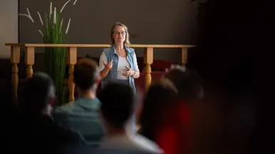 Een blonde bebrilde vrouw in wit t-shirt en jeansvestje spreekt een zaal toe. 