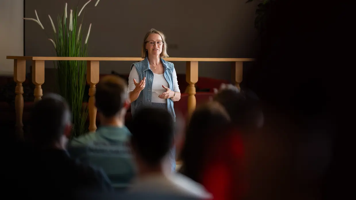 Een blonde bebrilde vrouw in wit t-shirt en jeansvestje spreekt een zaal toe. 