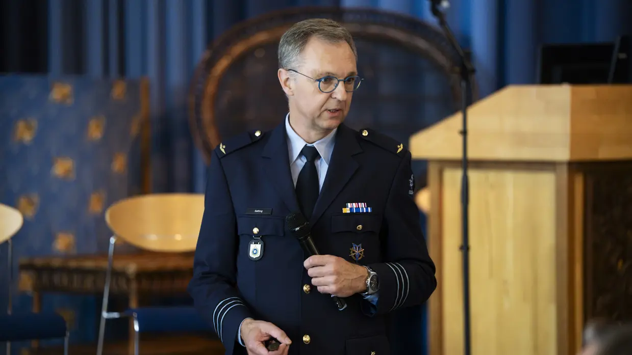 Een man met een bril, gekleed in een donkerblauw militair uniform met diverse onderscheidingstekens en een stropdas, houdt een microfoon vast terwijl hij spreekt. Hij staat voor een houten katheder in een ruimte met blauwe gordijnen op de achtergrond.