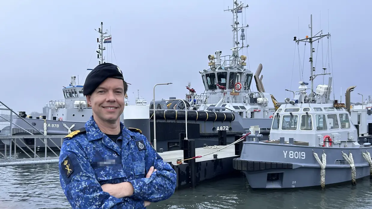 Een lid van de marine poseert voor 3 boten van de marine. 