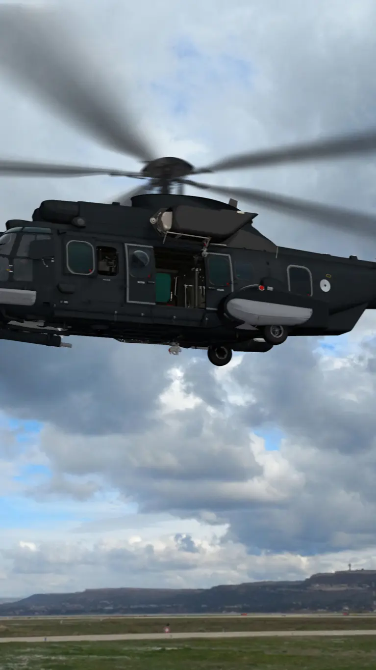 Een donkergrijze helikopter vliegt boven een vliegveld.