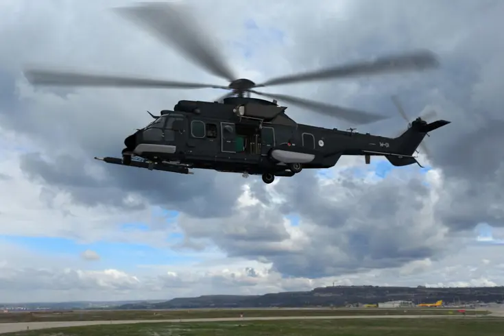 Een donkergrijze helikopter vliegt boven een vliegveld.