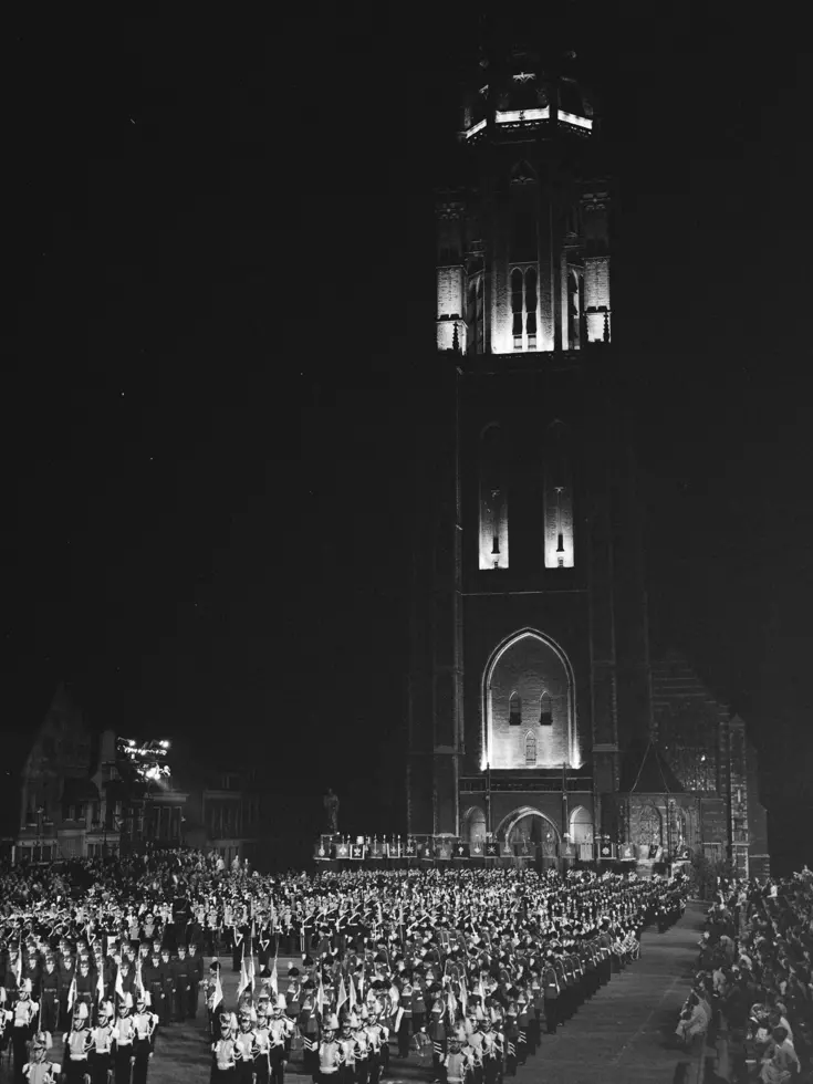 Een verlichte kerktoren met klok torent boven een menigte op een plein. Geüniformeerde muziekgroepen staan in formatie, alsof er een plechtige ceremonie of optocht plaatsvindt in het donker.