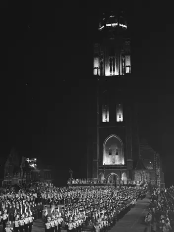 Een verlichte kerktoren met klok torent boven een menigte op een plein. Geüniformeerde muziekgroepen staan in formatie, alsof er een plechtige ceremonie of optocht plaatsvindt in het donker.