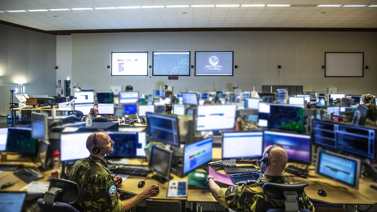 Overzichtsfoto van een ruimte met veel computerschermen. In de achtergrond op de muur hangen drie grote projectieschermen. Foto: