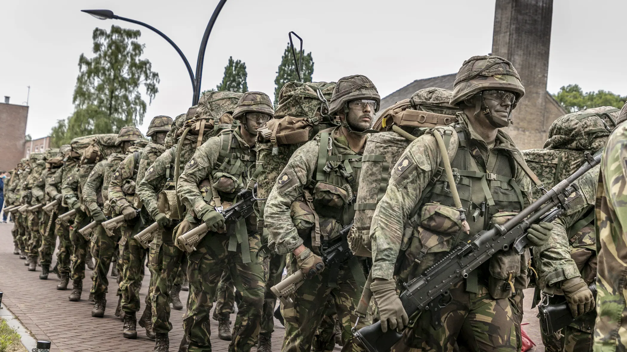 Militairen marcheren langs de fotograaf. Ze dragen een wapen, volle bepakking en hebben camouflage op het gezicht.