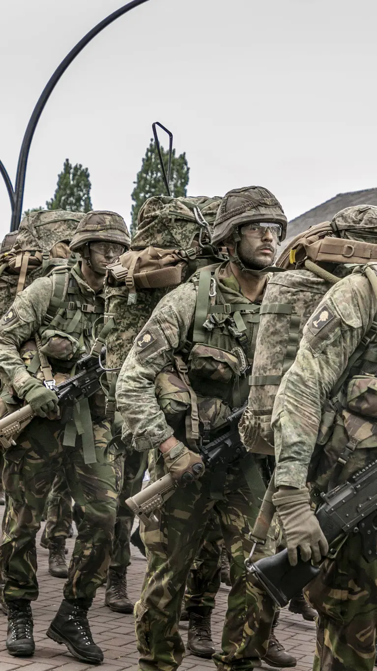 Militairen marcheren langs de fotograaf. Ze dragen een wapen, volle bepakking en hebben camouflage op het gezicht.