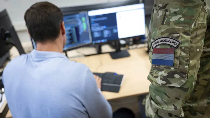 Een man zittend achter zijn computer, daarnaast een staande militair met op zijn arm de tekst ‘Korps Commandotroepen’.