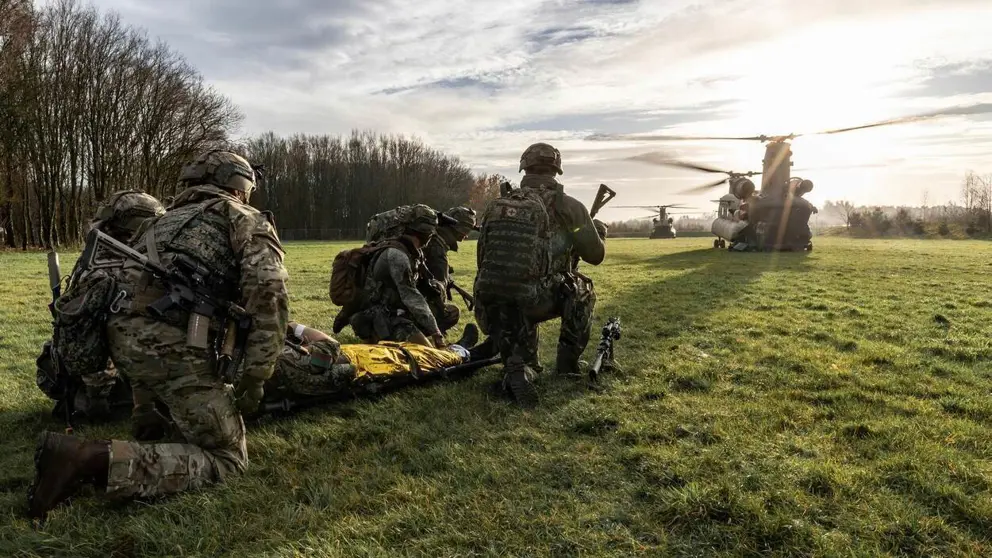 5 militairen zitten op hun knieën in het gras bij een gewonde op een brancard. 2 Chinook-transporthelikopters zijn zichtbaar op de achtergrond.